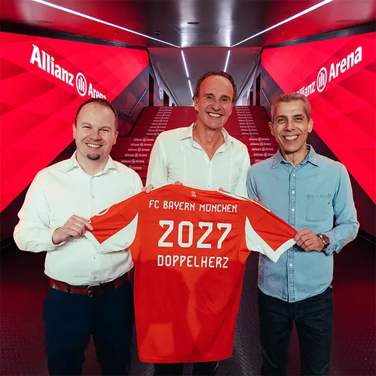 Trois hommes se tiennent ensemble dans un couloir de l'Allianz Arena. L'un d'eux tient un maillot du FC Bayern Munich, rouge, avec l'inscription "2027 DOPPELHERZ" au dos. L'ambiance est dynamique, soulignant un moment de célébration.