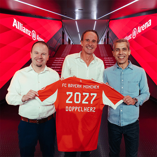 Trois hommes se tiennent ensemble dans un couloir de l'Allianz Arena. L'un d'eux tient un maillot du FC Bayern Munich, rouge, avec l'inscription "2027 DOPPELHERZ" au dos. L'ambiance est dynamique, soulignant un moment de célébration.