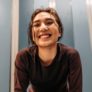 Une jeune femme souriante, vêtue d’un haut sombre, est penchée en avant. Ses cheveux sont humides et son regard exprime la joie et l’énergie. En arrière-plan, des murs de couleur bleue ajoutent à l’atmosphère dynamique.