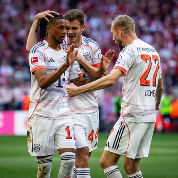 Des joueurs de football de l'équipe du Bayern Munich célèbrent un but. Un joueur est au centre, souriant et levant les mains, tandis que deux coéquipiers se rejoignent autour de lui, affichant des expressions de joie. L'ambiance est festive sur le terrain.