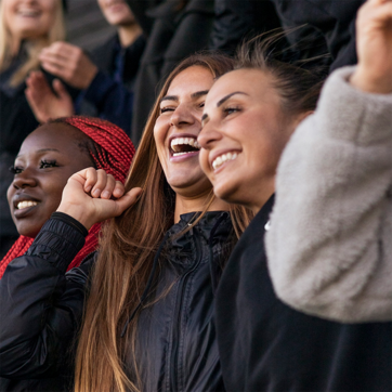 Trois femmes souriantes se tiennent côte à côte, exprimant leur joie. L'une d'elles a des cheveux longs et bruns, une autre porte des cheveux noirs avec des tresses rouges, et la troisième a des cheveux châtains et porte un manteau gris. Elles semblent célébrer un moment heureux.